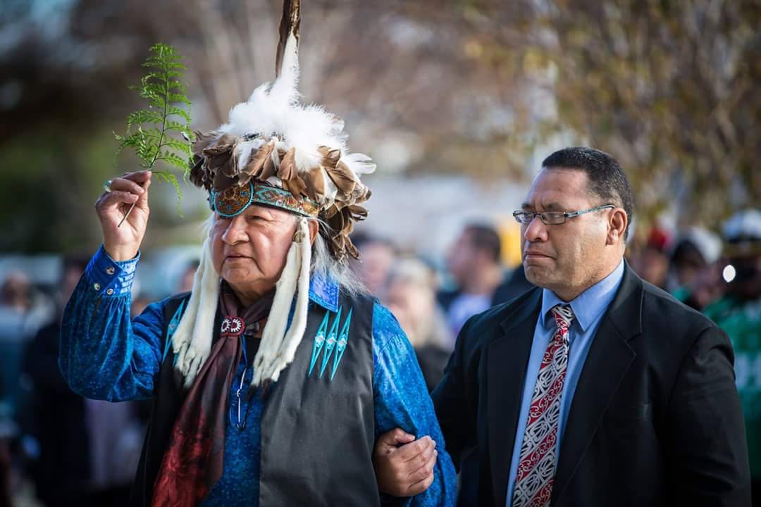 A kaumatua wearing a feather headdress raises a fern frond alongside a manuhiri during a powhiri